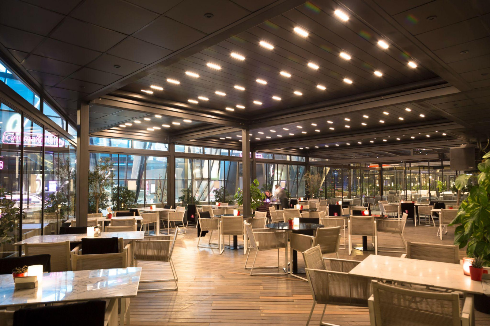 Açık havada yemek yemeye yönelik pergola çözümlerinin sergilendiği, cam duvarın dışında masa ve sandalyelerin bulunduğu bir restoran.