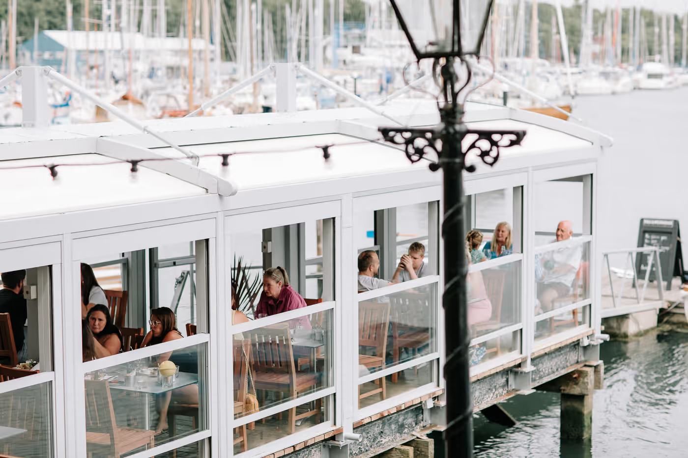 A view of people sitting at tables under a white pergola system and boats on the pier.