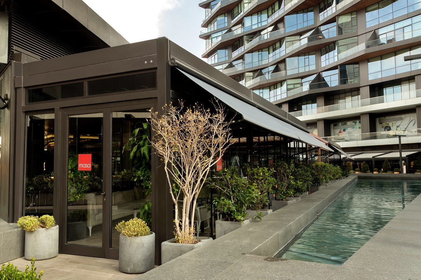 Önünde havuz ve yeşillikler bulunan, gölge ve konfor için Otomatik Tente Sistemlerinin ön plana çıktığı bir restoran.